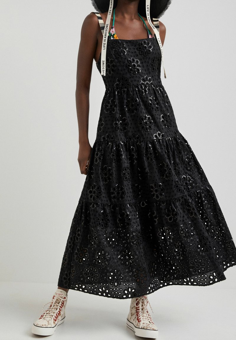 Robe maxi noire avec découpes florales, design à étages, bretelles spaghetti et détails complexes. Associée à des baskets blanches.