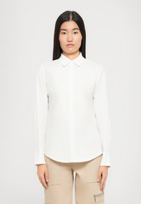 Donna asiatica con lunghi capelli lisci, che indossa una camicia bianca a maniche lunghe e pantaloni beige con tasche, in piedi davanti a uno sfondo neutro.