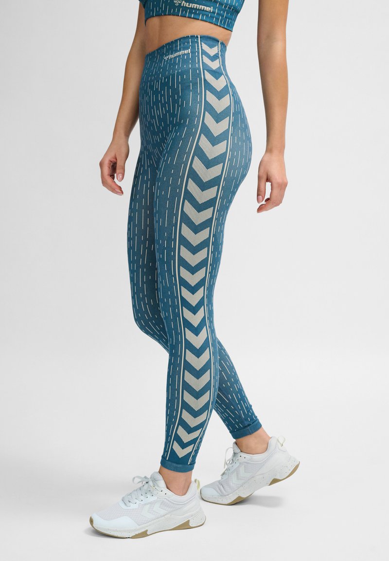 Blå leggings med vertikala linjer och ett chevronmönster på sidorna, tillverkade av ett stretchigt material, i kombination med vita träningsskor.