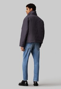 Giacca imbottita viola scuro con colletto alto, taglio leggermente oversize, abbinata a jeans azzurri chiari e calzature nere. Design semplice, senza motivi visibili.