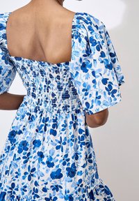 Robe à motif floral en bleu et blanc, avec des manches bouffantes, un dos smocké et une jupe à volants. Tissu doux avec une texture légère.