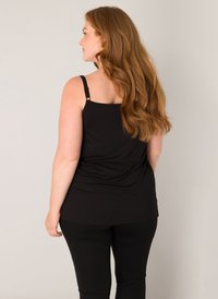 Vrouw met lang rood haar, gekleed in een zwart mouwloos topje en een zwarte broek, staand met haar rug naar de camera tegen een eenvoudige lichte achtergrond.