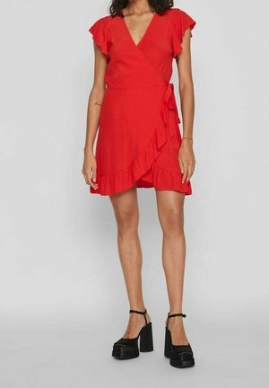 Rode wikkeljurk met korte gerimpelde mouwen en een zijstrik. Heeft een gerimpelde zoom en gladde stof. Gecombineerd met zwarte platformhakken.