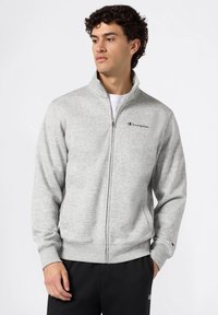 Sudadera de cremallera en gris claro, hecha de una tela suave y texturizada. Presenta un cuello alto y un logo bordado en el pecho. Puños de contraste.