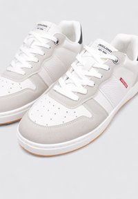 Baskets blanches et gris clair avec un design texturé, comportant des perforations, des lacets blancs et des semelles en caoutchouc. Logo de la marque visible sur la languette.