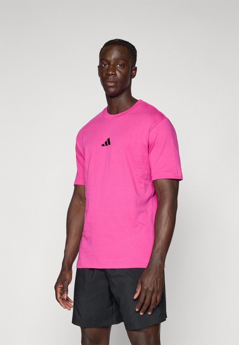 Fukszia pamut póló rövid ujjakkal és kerek nyakkivágással, fekete Adidas logóval a mellkason. A modellen fekete sort van.