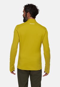 Maglia gialla a maniche lunghe con un motivo a trama, dotata di cuciture piatte e colletto alto. Logo del marchio "Mammut" ben visibile sul retro.