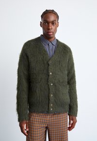 Πράσινο, φουντωτό κασκόλ με V-λαιμόκοψη, που διαθέτει έξι κουμπιά, δύο μπροστινές τσέπες και υφή. Φοριέται πάνω από ένα μπλε καρό πουκάμισο.