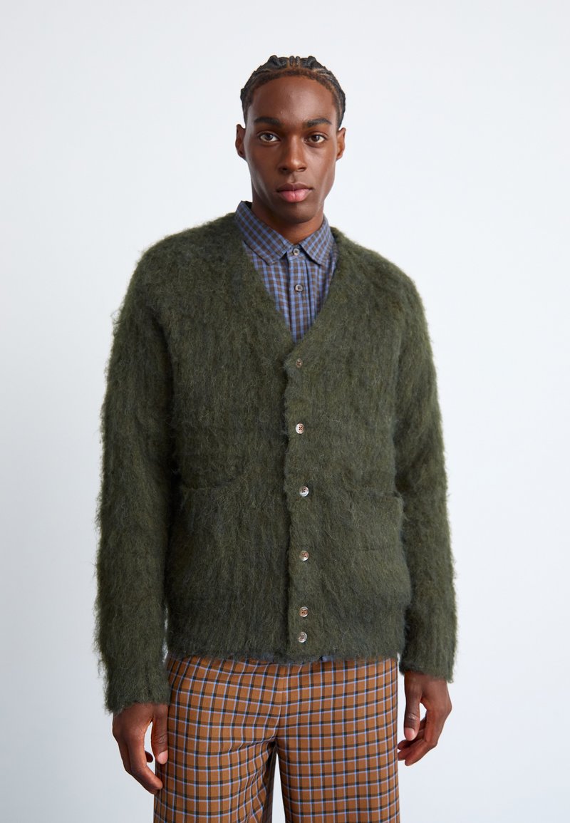 Πράσινο, φουντωτό κασκόλ με V-λαιμόκοψη, που διαθέτει έξι κουμπιά, δύο μπροστινές τσέπες και υφή. Φοριέται πάνω από ένα μπλε καρό πουκάμισο.