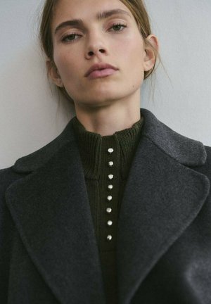 Abrigo de lana gris oscuro sobre una camiseta de punto acanalado verde oscuro que presenta un cuello alto y una fila vertical de seis pequeños botones plateados.