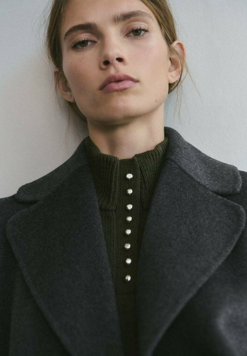 Cappotto di lana grigio scuro sopra un top a costine verde scuro con collo alto e fila verticale di sei piccoli bottoni argentati.