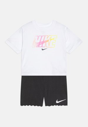 Biała koszulka z różowo-żółtym gradientem z grafiką "NIKE", w połączeniu z czarnymi szortami o falbaniastym wykończeniu i białym akcentem z logo.