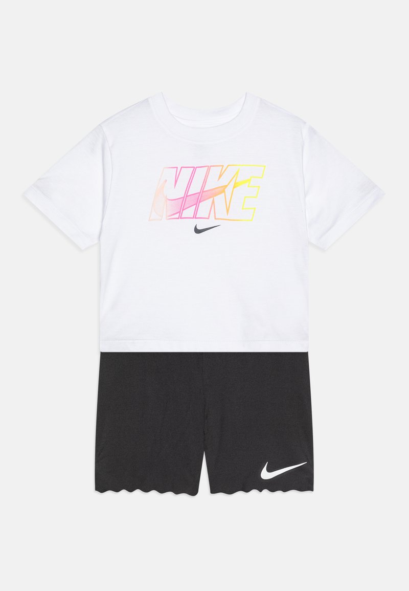 Balta T-kreklā ar rozā un dzeltenu gradientu "NIKE" grafiku, apvienota ar melniem īsiem šortiem ar skalotu apakšmalu un baltu logotipa akcentu.
