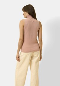 Kobieta w bezrękawniku w kolorze przykurzonego różu i szerokich kremowych spodniach, stojąca plecami do aparatu.