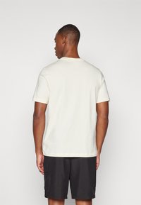 Tricou bej deschis din bumbac, cu guler rotund și mâneci scurte, purtat cu pantaloni scurți închiși la culoare. Design curat, simplu.
