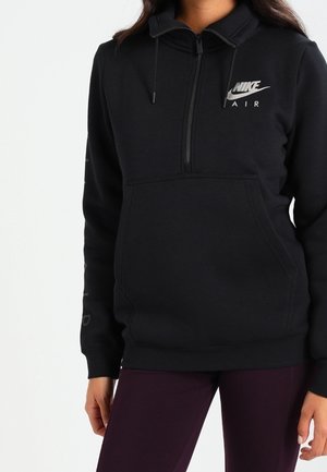 Persoon draagt een zwarte Nike Air half-zip hoodie met een voorzak en bordeauxrode broek, staand tegen een effen witte achtergrond.