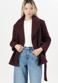 Bordeaux uldblandet frakk med sjalskrave, frontknude og lange ærmer, parret med højhævede blå jeans og en hvid top.