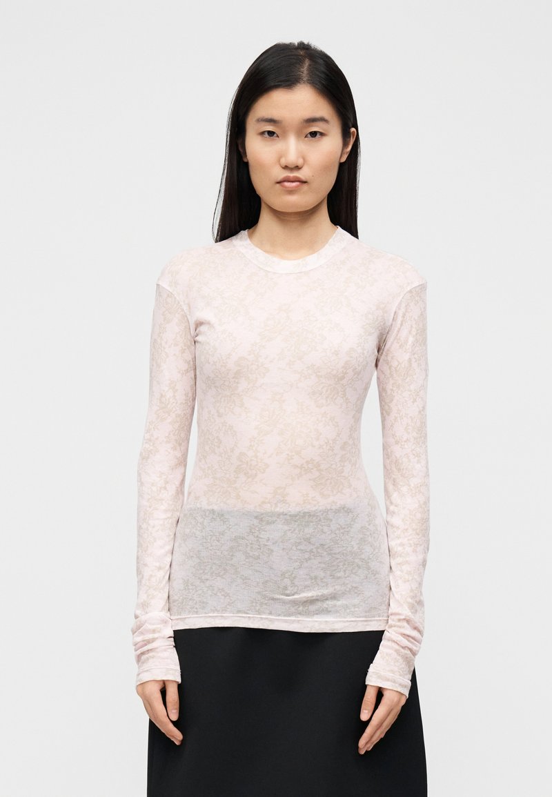 Dolga majica svetlo roza barve z rožnatim vzorcem, prosojne teksture, z okroglim izrezom in oprijetim krojem; nošena preko temnejše krila.