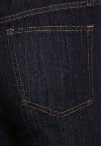 Jeans de mezclilla oscura que presentan un bolsillo trasero con costuras naranjas en contraste y una textura ligeramente desgastada. Los detalles de las costuras son visibles.