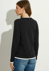 Schwarzer Pullover mit langen Ärmeln und rundem Ausschnitt, verziert mit einem cremefarbenen Saum am Kragen und am Saum. Getragen über blauen Jeans.