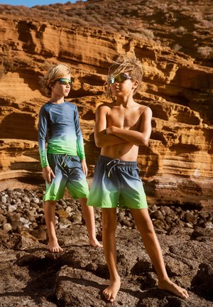 Pojkarna har på sig gradientbadkläder; en i långärmad tröja i blågrön och matchande shorts, den andra i shorts med gröna byxor. Båda har solglasögon på sig.