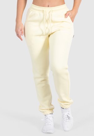 Woman wearing cream-colored sweatpants with elastic ankles and white sneakers, standing against a plain light background.