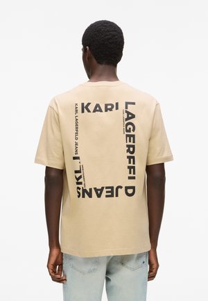 Homme portant un t-shirt beige avec le texte noir "KARL LAGERFELD JEANS" disposé dans un cadre rectangulaire au dos, associé à un jean bleu clair.