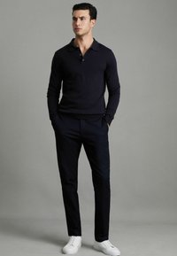 Polo cu mâneci lungi de culoare bleu marin, textură netedă, cu sistem de închidere cu trei butoane. Combinat cu pantaloni negri și adidași albi, stând cu mâinile în buzunare.