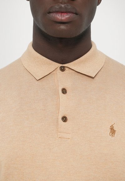 bézs színű, texturált anyagból készült galléros póló, klasszikus gallérral, három barna gombbal és egy kis hímzett logóval a bal mellrészen.