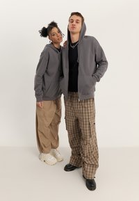 Due persone stanno fianco a fianco. Una indossa una felpa grigia con zip e pantaloni cargo. L'altra indossa una maglietta nera e pantaloni a quadri.