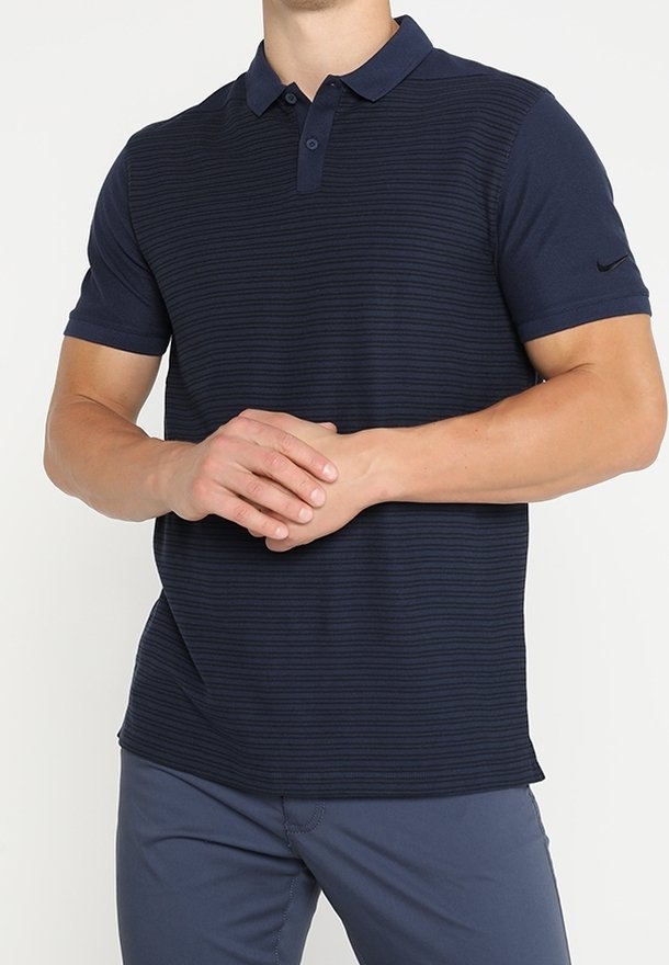 Homme portant un polo à manches courtes bleu marine avec des rayures horizontales subtiles, les mains jointes devant à hauteur de la taille.