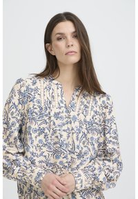 Blouse florale en tissu crème avec des imprimés botaniques bleus. Caractéristiques : encolure en V, plis et poignets élastiqués pour un look ajusté.