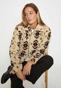 Krēma un brūna rakstota flīsa jaka ar rāvējslēdzēja aizdari un apkakli, kurai ir teksturēts dizains un brīvs piegriezums.