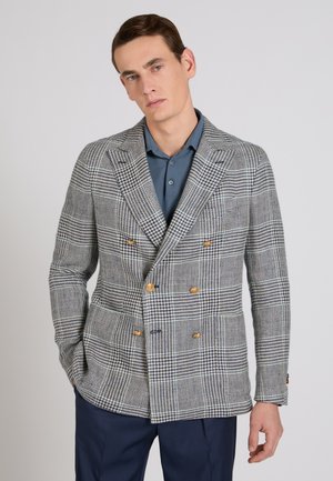 Mężczyzna w szaro-białej dwurzędowej marynarce w kratę z złotymi guzikami, na niebieskiej koszuli z kołnierzykiem, stojący z jedną ręką w kieszeni.