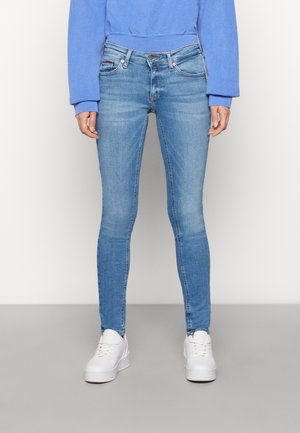 Henkilö pukeutuneena vaaleansinisiin skinny-farkkuihin, siniseen collegepaitaan ja valkoisiin korollisiin lenkkareihin, seisomassa yksinkertaisen harmaan ja valkoisen taustan edessä.