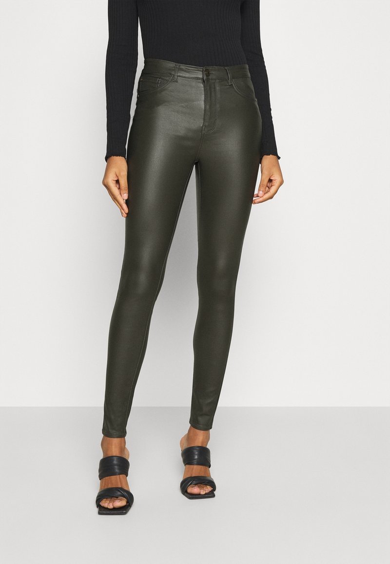 Femme portant un pantalon skinny taille haute en cuir vert olive, un top noir à manches longues côtelé et des sandales noires à bouts ouverts.