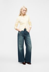 Ξανθιά γυναίκα στέκεται με τα χέρια στις τσέπες φορώντας ανοιχτό κίτρινο κουμπωμένο πουλόβερ, φαρδιά ξεθωριασμένα μπλε τζιν και σκούρα καφέ παπούτσια σε λευκό φόντο.