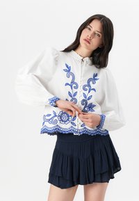 Blusa bianca con ricami floreali blu e polsini arricciati, abbinata a una mini gonna plissettata scura. I dettagli principali includono una chiusura con bottoni e un orlo ondulato.