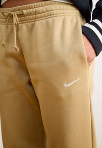 Beżowe spodnie dresowe wykonane z miękkiego materiału, z elastycznym paskiem w talii i sznurkiem. Na udzie znajduje się małe białe logo Nike.
