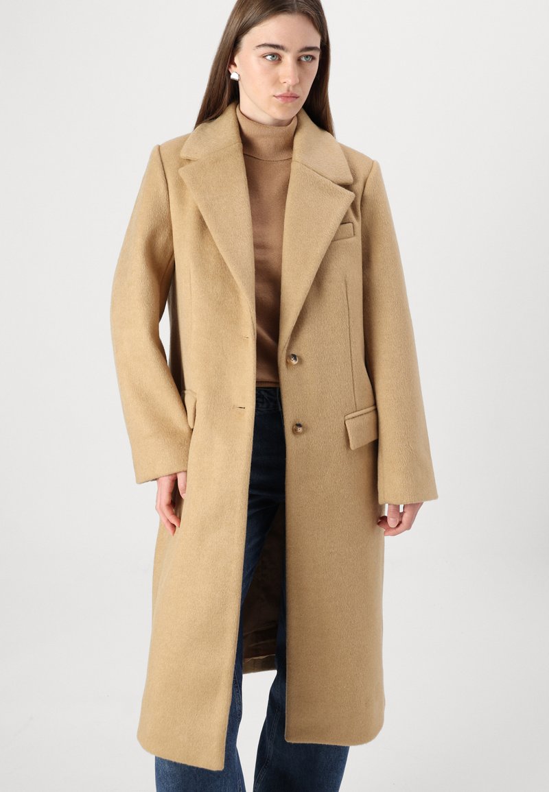Beżowy wełniany płaszcz z dużymi klapami, dwoma guzikami z przodu i kieszeniami bocznymi. Noszony na brązowy golf i zestawiony z ciemnymi dżinsami.