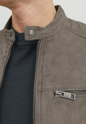Gros plan d'une personne portant une veste en suède grise avec un col à bouton-pression et une poche zippée par-dessus un pull en tricot foncé.