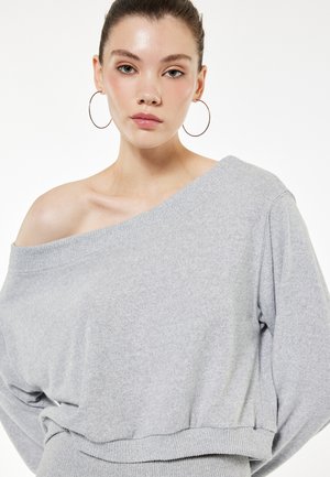 Jonge vrouw met naar achteren getrokken haar, draagt grote creolen en een grijze off-shoulder trui, poserend tegen een effen witte achtergrond.