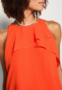 Haut sans manches orange en tissu léger avec un détail à volants superposés au niveau de l'encolure. Des perles sont portées en tant que collier.