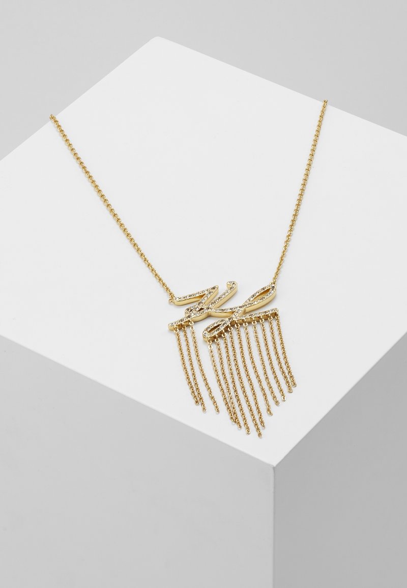 Collier en chaîne dorée avec un pendentif scintillant "KL" et plusieurs pampilles en chaîne dorée, présenté sur une surface géométrique blanche.
