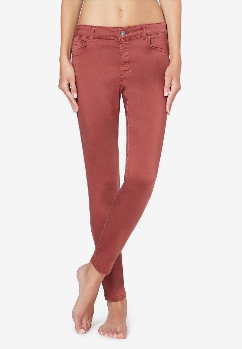 Jean slim couleur rouille porté par une personne pieds nus, debout, jambes croisées aux chevilles sur fond blanc.