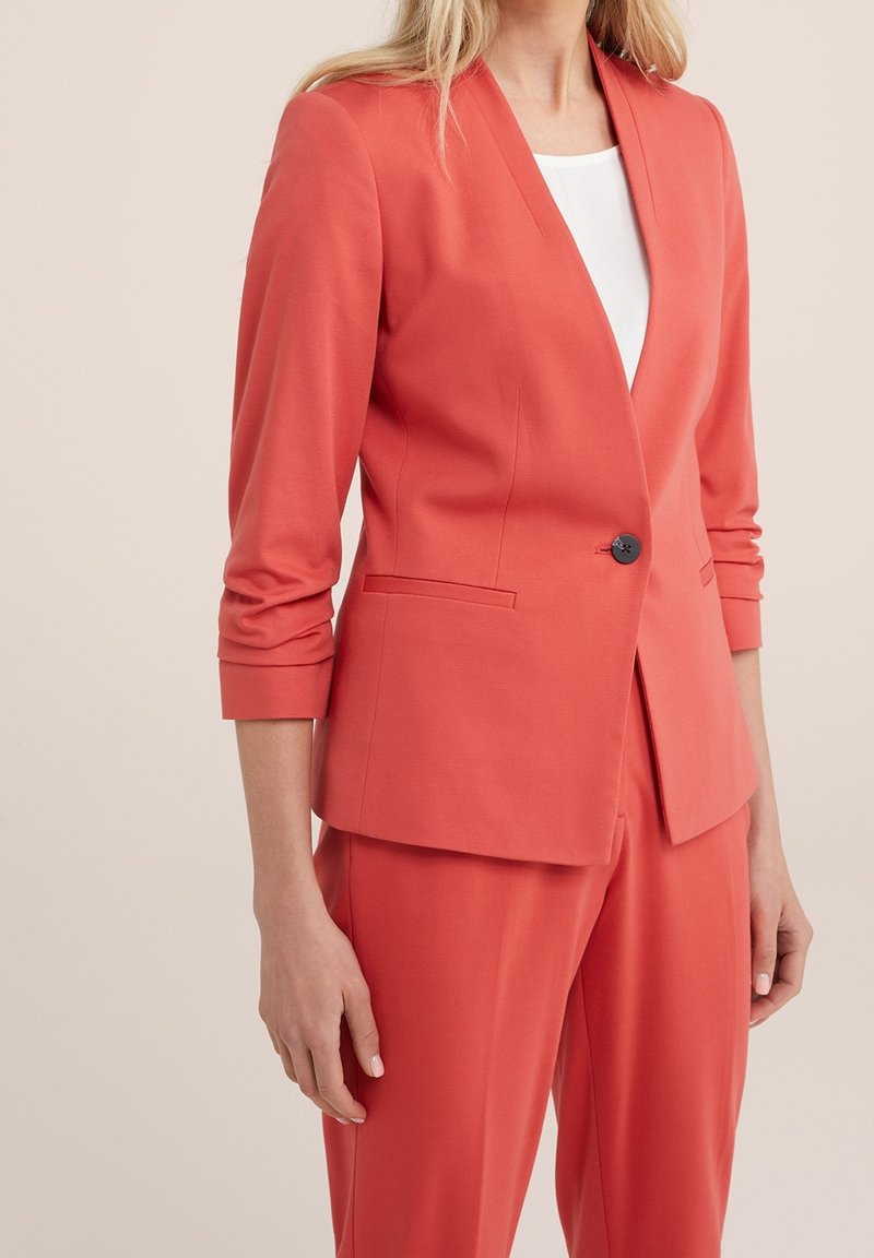 Korálový blazer s výstřihem do V, zapínáním na jeden knoflík, dlouhými rukávy a odořeným střihem. Hladká látka, postranní kapsy a minimalistický design.
