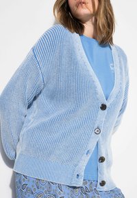 Vaaleansininen neulecardigan, jossa on V-pääntie, neljä tummaa nappia ja ribattu helma. Yllään yhteensopiva sininen t-paita ja kuvioidut housut.