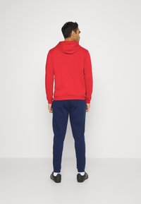 Roter Kapuzenpullover mit Kängurutaschen, kombiniert mit marineblauen Jogginghosen, die an den Knöcheln umgeschlagen sind. Der Stoff wirkt weich und komfortabel.