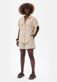 Beige linen short-sleeve shirt with two pockets, paired with matching shorts. Features a relaxed fit and brown platform sandals.