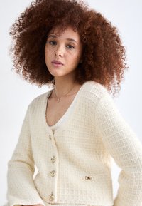 Cremefarvet cardigan med V-udskæring, fem guldknapper og en guldsløjfeaccent på venstre side. Blødt strikkemateriale.
